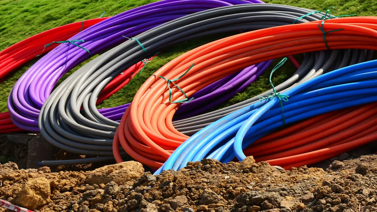 Brightly colored coils of fiber-optic conduit staged on fresh earth beside an open trench at a broadband construction site.