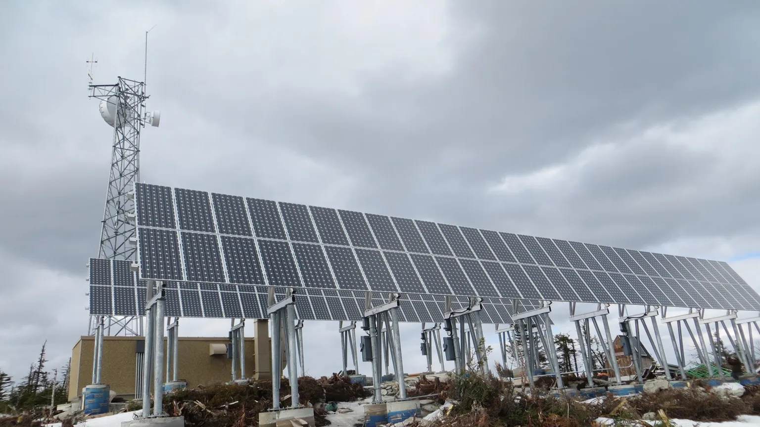 A solar-powered remote LMR communications site — solar panel array on a structural framework with a comms tower in the background under an overcast mountain sky.