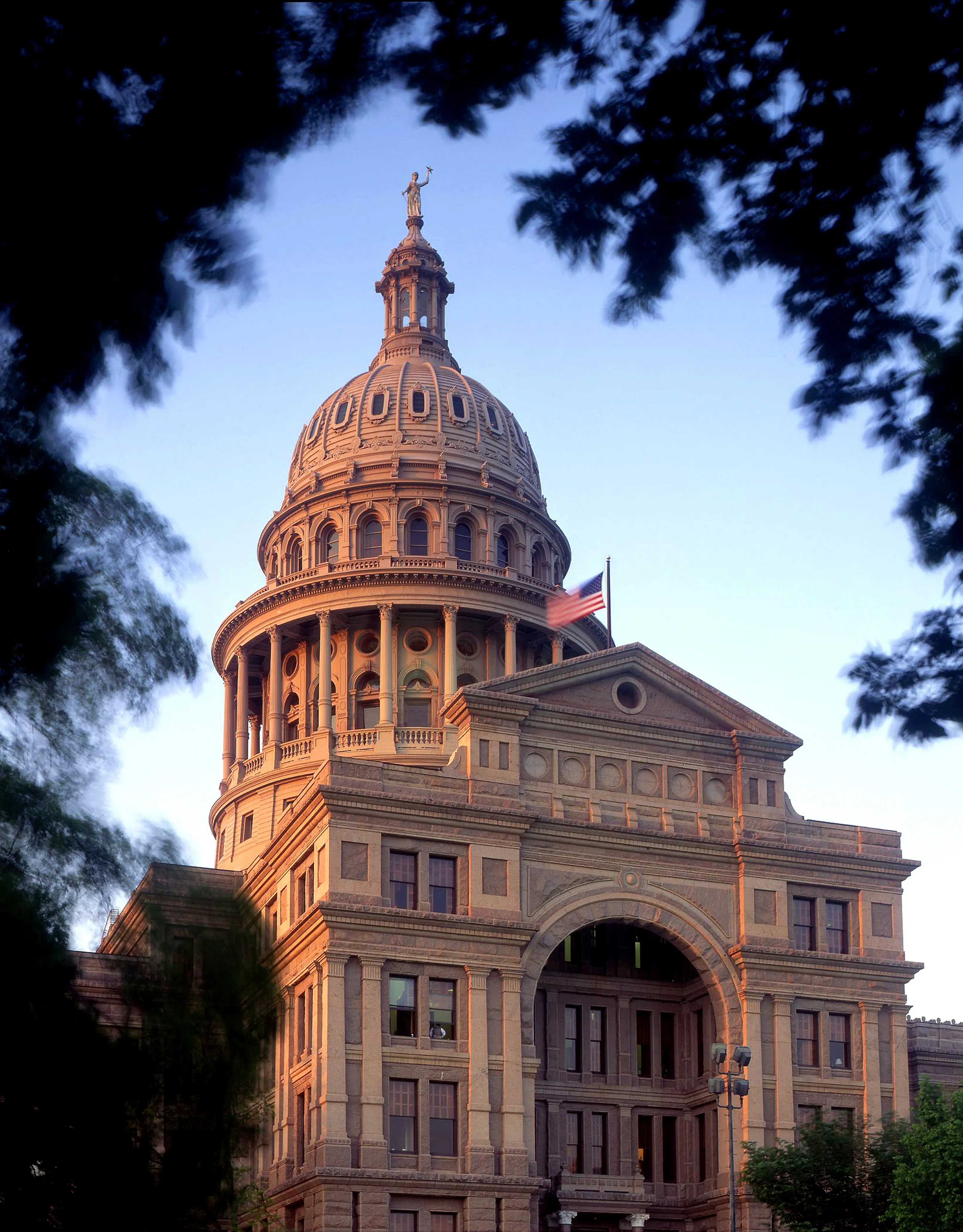 The Texas State Capitol dome in Austin at golden hour, framed by oak leaves.