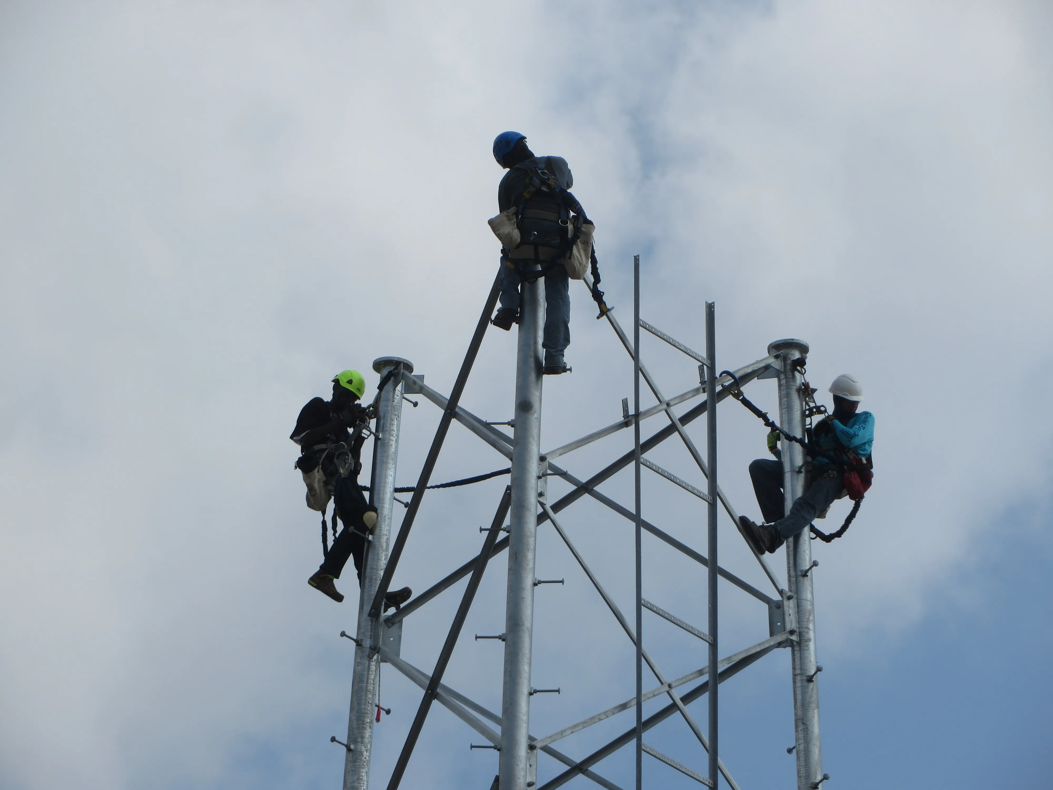 Three workers in safety harnesses climbing a self-support telecommunications tower against a partly cloudy sky.